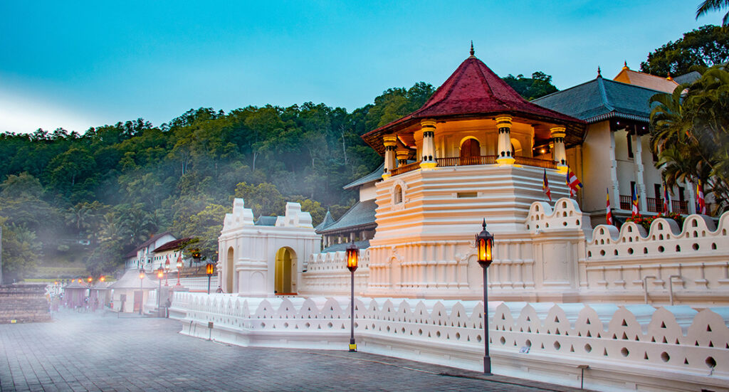 Temple of the Sacred Tooth Relic in Kandy lit up at dusk with forested hills behind